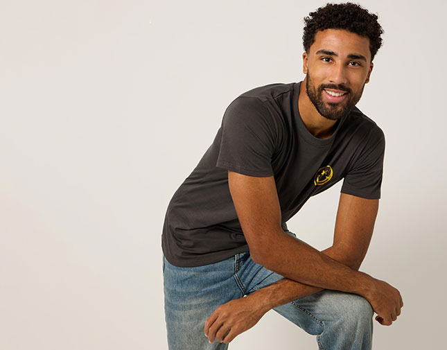 Male model wearing a dark charcoal T-shirt with a yellow smiley face chest graphic and light blue jeans, posing against a plain light background.