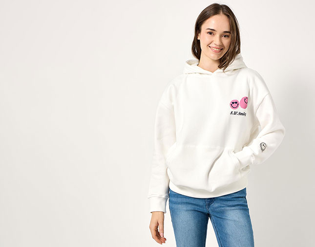 Female model wearing a white hoodie with pink smiley graphics, &ldquo;S.W. Smiley&rdquo; text, and blue jeans against a plain light background.