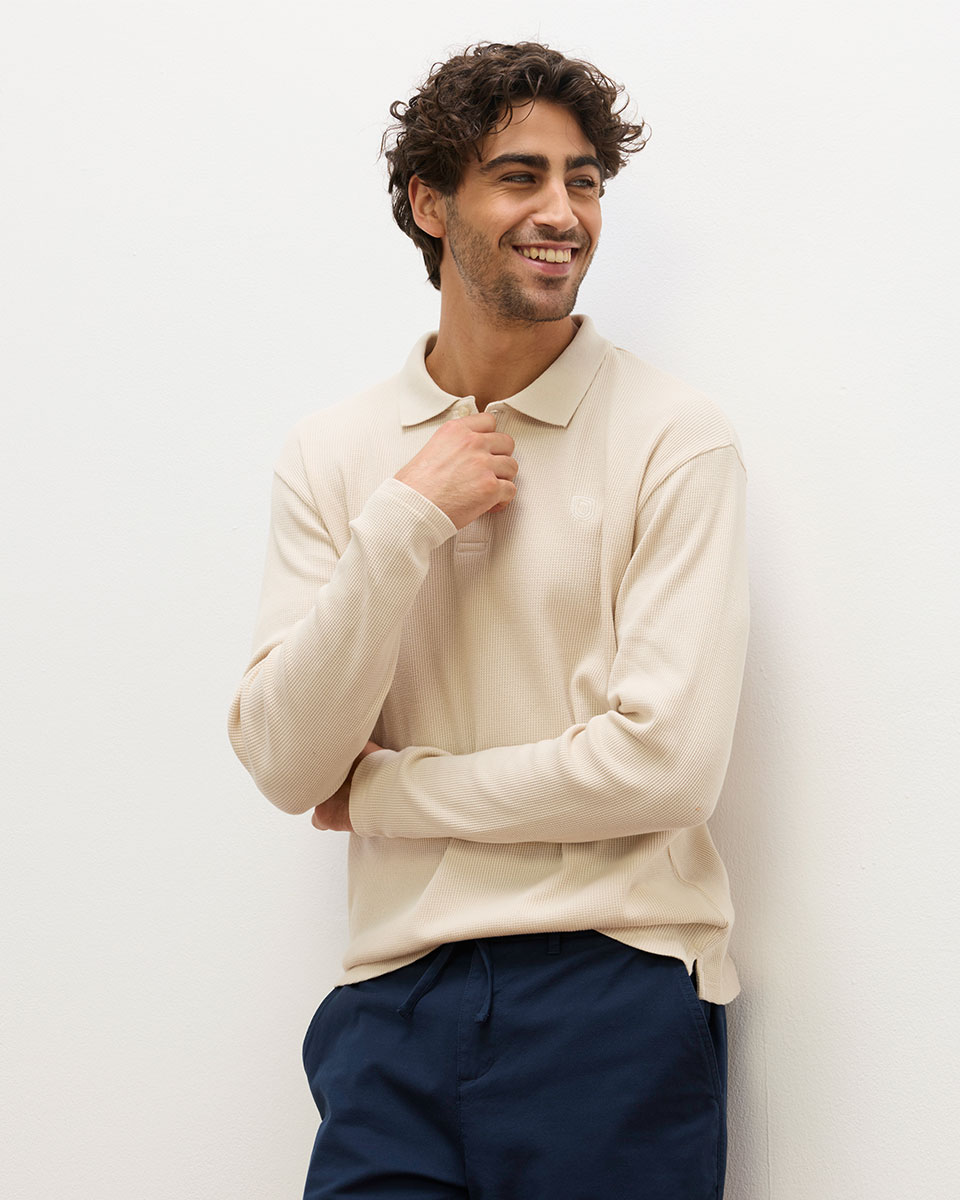 Smiling man leaning against a wall, wearing a cream textured knit polo shirt with a small chest logo and navy drawstring trousers.